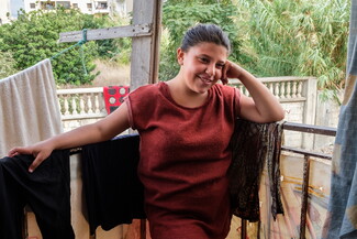 A young girl leans on a balcony railing, on which laundry dries. Her hair is neatly pushed back and she looks shyly away.