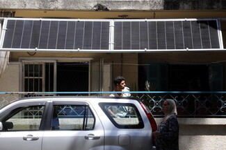 A man stands on a ground-floor balcony adjacent to the road, as solar panels hang above him. Only a few meters in front, a woman walks by a parked car.