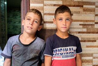 Two little boys with striking blue eyes stand side by side by the front of their house.
