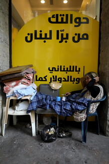An old woman sits on a makeshift bed she has made in front of OMT. She is lying across two plastic chairs and is covered with a blue blanket.