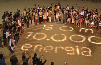 A large group of people circle around writing in the sand, lit by candles, that reads: "Som 10,000 energia."