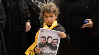  A child, draped in a Hezbollah scarf, carries a flyer honoring the martyred Hussein family—Hassan Hussein, his wife Ruwayda Mustafa Hussein, and their son Ali Hussein—during the funeral procession. March 6, 2024. Houla, South Lebanon. (Fatima Joumaa/The Public Source)