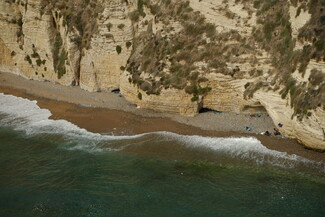 A small sandy beach lies at the base of steep, rugged cliffs with sparse vegetation. Waves gently wash ashore, and a few people and items are visible near the cliffs.