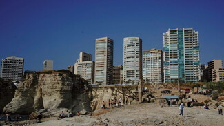  A landscape view features residential towers in the background. In the center of the image and to the right, there are temporary structures made of wood, corrugated metal, and plastic. A couple of feluccas, a few motorcycles, and a car are parked next to them. Young men are gathered in different spots, some by the water, others on the cascading plateau.