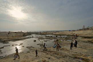 As the sun begins to set, people at Dalieh start gathering their belongings and children, preparing to leave. Some linger in the water, while others stand behind them..
