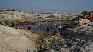Parents sit on rocks overlooking the shallow pool at Dalieh while kids swim. The area is surrounded by trash such as plastic bottles and chip bags, stuck in the rocks