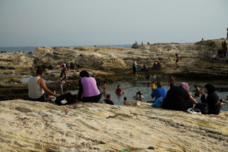 A group of people sit on a cliff overlooking the water while others swim in the background.