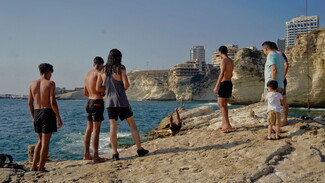 Around five people stand on a cliff waiting to dive into the water. 