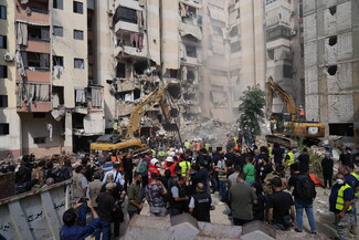 A bulldozer and a crowd of people stand near and atop a large mound of rubble, in the middle of a residential area, adjacent to the destruction and ruin of nearby buildings.