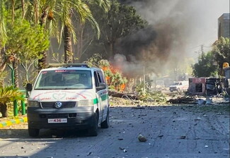An ambulance on a debris-strewn street with smoke and flames visible in the background. Trees and damaged vehicles are present.