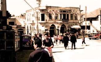 A lively street scene in front of a building with arched windows and a sign in Arabic and French. People are walking, and cars are parked nearby.