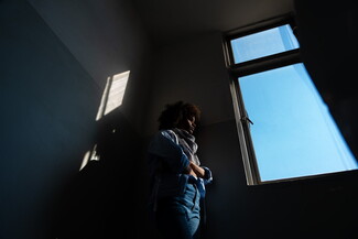 A photo taken from below; a woman stands by a window in a dark room. The sky outside is a clear blue. The sunlight hits her softly.