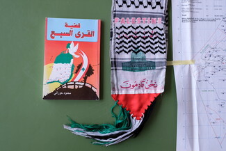 A book titled “The Case of the Seven Villages” to the left of a keffiyeh with a print of Al Aqsa Mosque that reads: "PALESTINE" and "We are coming."