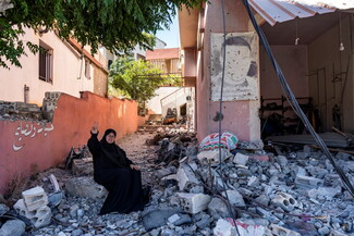 An elderly woman, wearing a black hijab, sits on the rubble of a destroyed home, with a partially standing structure in the background, in a village in South Lebanon.