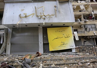 A bright yellow poster hangs among rubble and a shuttered storefront. The poster, written in Arabic, reads: “In the name of God, we will certainly be victorious,” and is signed “Nasrallah.”