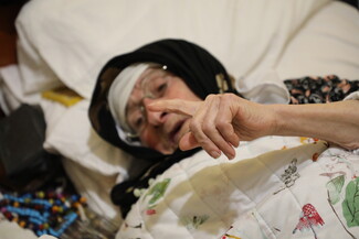 An elderly woman in her bed, raises her hand.
