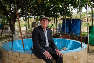 An older man wearing a cap sits on the edge of an emptied out pool, surrounded by trees.