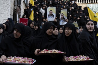 Three women in black carry baskets full of rose petals at a funeral. In the background are other funeral-goers, carrying photos of the martyred and flags.