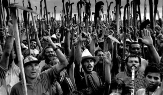 A black-and-white photo shows Brazilian farmers from the Landless Workers Movement raising their fists and holding shovels and agricultural tools high during a demonstration, their tools symbolizing both their labor and their struggle for land rights.
