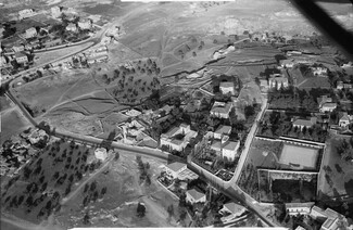 Aerial view of a mid-20th century village with scattered houses, open fields, trees, and a winding road.
