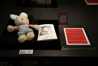A small, worn rabbit toy rests on a display surface beside a pamphlet detailing the story of a child victim of cluster bombs.