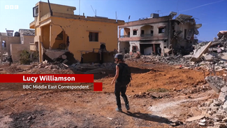 A woman stands amid rubble, center frame. Around her are fully demolished and semi-demolished buildings. She is wearing a helmet and flak jacket. To her left is a graphic that reads: “Lucy Williamson | BBC Middle East Correspondent”