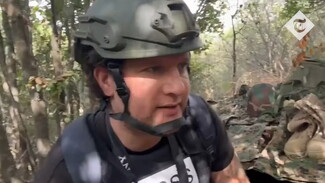 A still image from a video. A man in a helmet and press vest crouches in a lush, green area. Behind him appears to be military apparel.