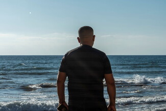 A man stands with his back to the camera and the sea before him. The sun is bright and the water is a deep blue. 