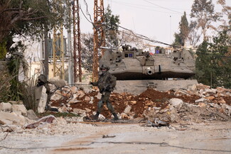 A military tank positioned behind a large pile of rubble. A soldier walks in front of the tank and looks right into the camera.