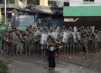 A female demonstrator confronts a line of uniformed military personnel holding shields and firearms stand in front of a damaged building. The scene is tense and guarded.