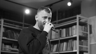 A bearded man passionately sings into a microphone, set against a backdrop of bookshelves. The black and white image conveys an intimate, artistic atmosphere.