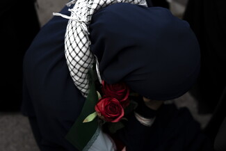Dressed in black, a woman sits on the floor, an Iranian flag wrapped around her arm, a kuffiyeh wrapped around her neck, and roses on her chest.