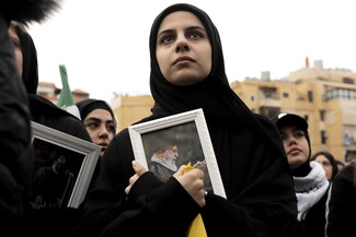 A woman holds a framed photograph of the late Ayatollah praying, pressing the image to her chest with one hand. She tightly holds a yellow Hezbollah sash with the other, her gaze fixed forward as she listens to the chanters.