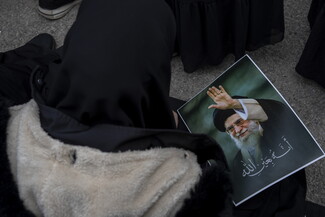 A woman wearing a black head scarf and a thick winter jacket faces away from the camera and is crouched on the ground. She bows her head. In her lap is a photograph of the late Ayatollah Khamenei.