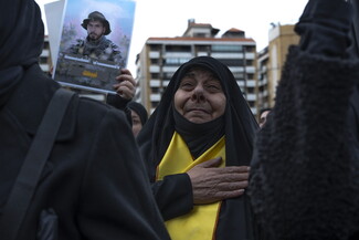 Clad in black, an elderly woman weeps as she beats her chest with her right hand, a Hezbollah sash wrapped around her shoulders. To the left of her, a hand lifts a photograph of the fighter Hassan Hamid (Abu Turab), marked with the words: “With my brother's martyrdom, I am at your service.”