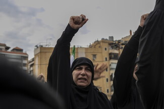 A woman clad in black raises her fist to the sky and chants among a crowd. A digital tasabih is visible on her finger.
