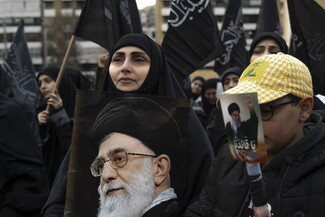 A woman looks onward as she carries a portrait of the martyred Supreme Leader close to her chest, resting her chin on it. Her son stands beside her, wearing a yellow Hezbollah cap and carrying a poster of Khamenei raising his fist, with the words “Oh our leader!” printed on it. Behind them, women raise black banners inscribed with “Oh Zainab the Great.”