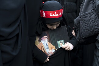 A young girl clutches a photograph of Imam Khamenei seated by a portrait of Imam Khomeini. A red headband from Imam Reza’s shrine adorns her head. It reads: “Sons of Fatima.”