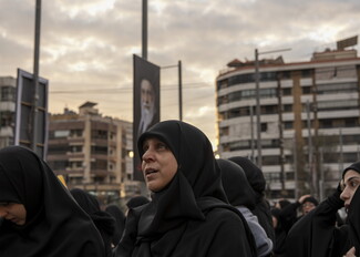 Gaze to the sky, a woman clad in black prays among a crowd of other women clad in black. A large poster of the martyred Ayatollah Khamenei is raised high on a pole behind her.