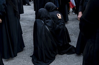 Among a crowd of women dressed all in black, two sit crouched on the ground. The woman on the left has her hand on the back of the woman on the right, soothing her.