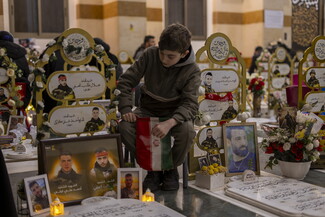 Holding the Iranian flag, a young boy sits in silence beside the graves of martyred resistance fighters Jalal Taleb al-Affi “Mohammad Ali” and Hajj Ahmad Ali Karaki “Jihad” at the Rawdat al-Hawraa Zainab military cemetery. Dahieh, Lebanon. March 1, 2026. (Fatima Joumaa/The Public Source)