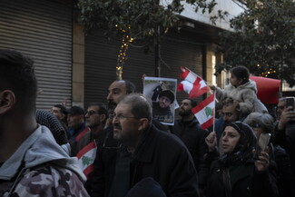 Two little girls held up by their fathers; they sit on their shoulders and carry a Lebanese flag and two books. The book on the left has a yellow cover and a photo of the late former Secretary General of Hezbollah, Sayyed Hassan Nasrallah. The book on the right has a green cover and a photo of the late Imam Musa al-Sadr.