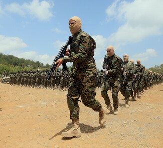 Armed soldiers in camouflage and tan balaclavas march in formation across a dirt field. A cloudy sky and forested background convey a tense atmosphere.