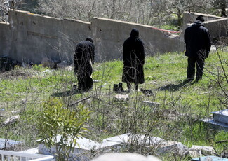 Three people in dark clothing stand on a grassy field near a damaged concrete wall, facing away. The scene feels somber and desolate.