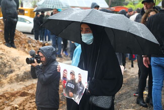 A woman stands in a mass cemetery under an umbrella, holding a photo of slain journalists Ali Shoeib, Fatima Ftouni, and Mohammad Ftouni.