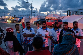 A group of people, some in matching shirts, stand and talk near tents and vehicles at dusk, with others gathered around them.