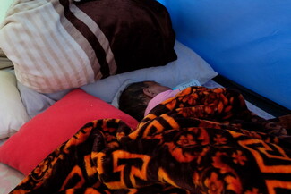 A baby sleeps on pillows inside a tent, covered with a patterned brown blanket and surrounded by cushions.
