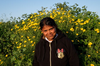 A woman in a black jacket stands against a background of greenery and yellow flowers.