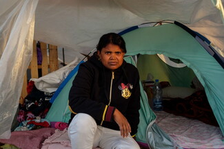 A woman sits at the entrance of a small, green tent, surrounded by blankets and personal items. 