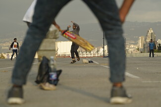A group of young men play cricket in an open, paved area against a cityscape backdrop. The focus is on a player mid-swing, conveying excitement and energy. We see the swinging player framed through the legs of the catcher.